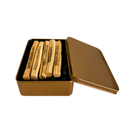 Five wooden Ecuadorian Palo Santo sticks laser engraved with viral social media phrases "PLOT TWIST VETERAN", "BOUNCE-BACK BADDIE", "PIVOT PRINCESS", "REINVENTION QUEEN", and "I AM THE PLOT TWIST" arranged in a square tin filled with black shredded paper on a white background.