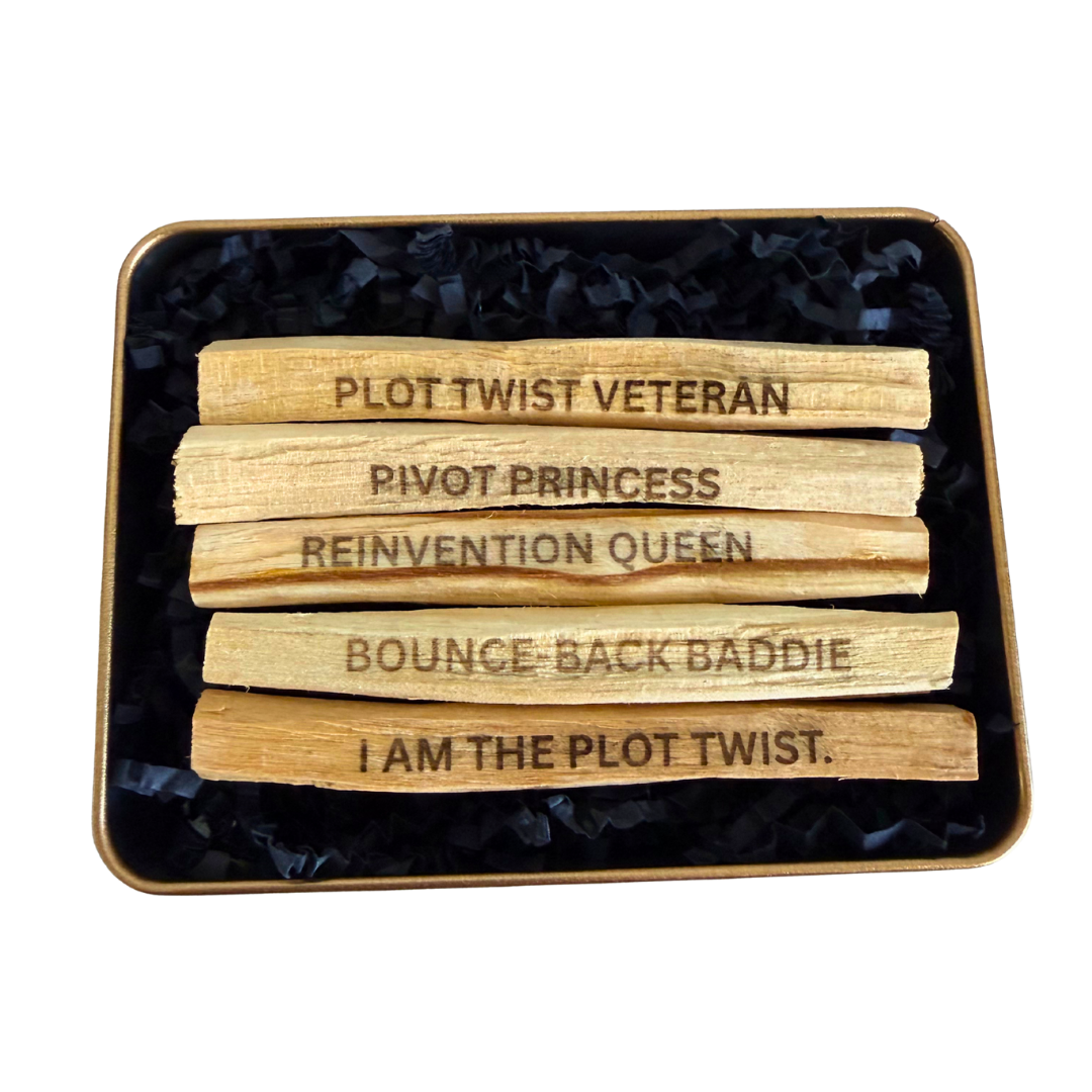 Five wooden Ecuadorian Palo Santo sticks laser engraved with viral social media phrases "PLOT TWIST VETERAN", "BOUNCE-BACK BADDIE", "PIVOT PRINCESS", "REINVENTION QUEEN", and "I AM THE PLOT TWIST" arranged in a square tin filled with black shredded paper on a white background.