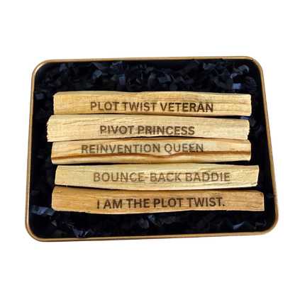 Five wooden Ecuadorian Palo Santo sticks laser engraved with viral social media phrases "PLOT TWIST VETERAN", "BOUNCE-BACK BADDIE", "PIVOT PRINCESS", "REINVENTION QUEEN", and "I AM THE PLOT TWIST" arranged in a square tin filled with black shredded paper on a white background.