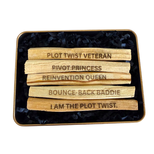 Five wooden Ecuadorian Palo Santo sticks laser engraved with viral social media phrases "PLOT TWIST VETERAN", "BOUNCE-BACK BADDIE", "PIVOT PRINCESS", "REINVENTION QUEEN", and "I AM THE PLOT TWIST" arranged in a square tin filled with black shredded paper on a white background.